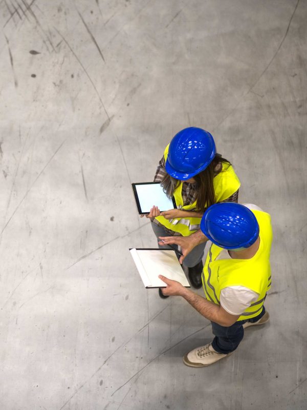 Top view of two industrial workers wearing hardhats and reflective jackets holding tablet and checklist on gray concrete background.